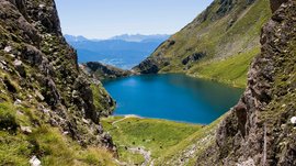 The Altfasstal valley, Meransen The image shows a deep blue mountain lake, surrounded by steep, green mountain slopes. In the background, there are more mountain ranges and a clear blue sky, conveying a calm and majestic landscape.