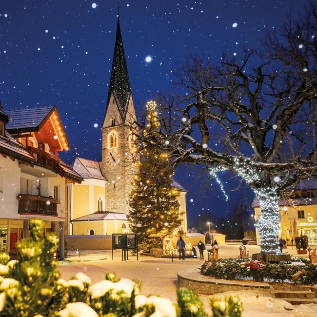 Tearna Advent Verschneiter Weihnachtsplatz mit beleuchtetem Tannenbaum und Kirchturm bei Nacht