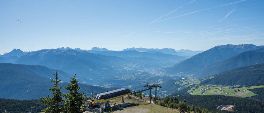 The Gitschberg webcams The picture shows a vast mountain landscape under a clear blue sky. In the foreground, there is a cable car station on a mountain peak, and in the background, several mountain ranges as well as a valley with scattered villages and forests can be seen. Two paragliders are flying high in the sky above the scene.