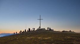 Vals - Jochtal - Steinermandl The picture shows a group of people gathered around a large summit cross on a peak. The sky is clear and shines in gentle blue and orange tones, indicating sunrise or sunset.