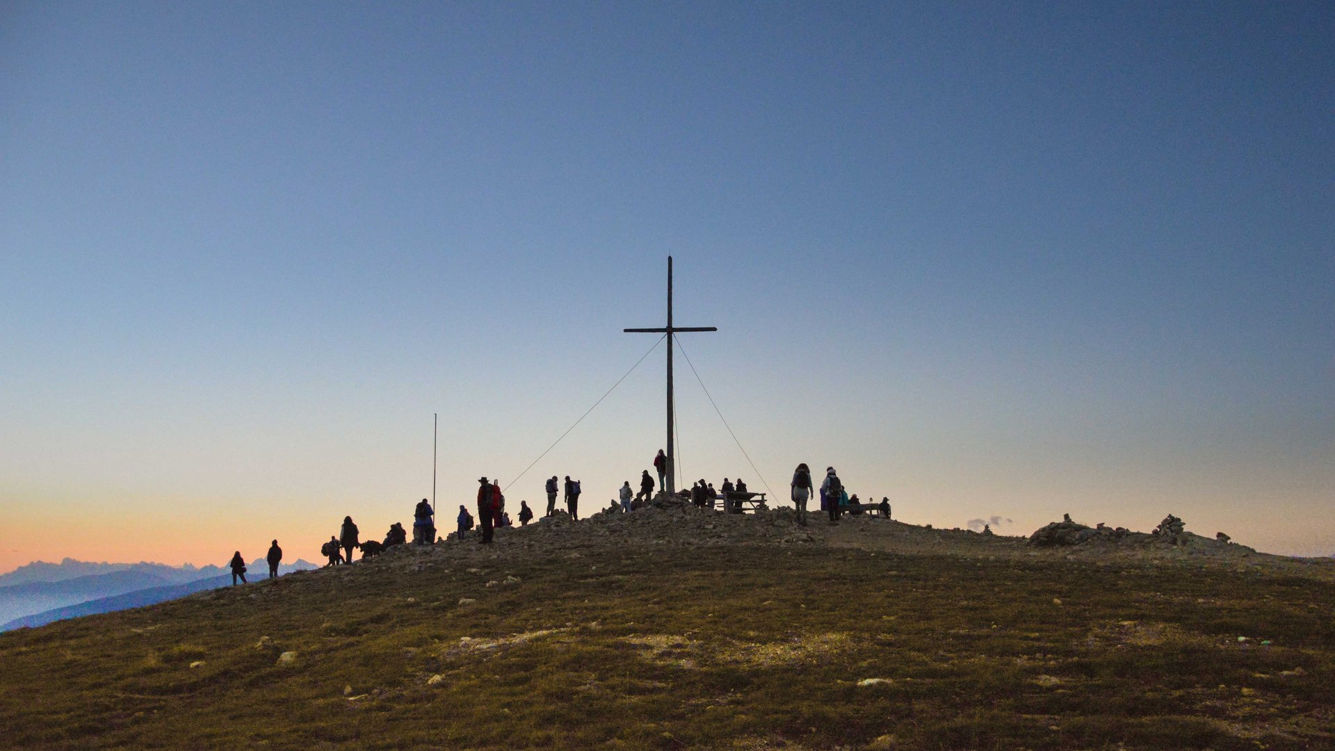 Vals - Jochtal - Steinermandl The picture shows a group of people gathered around a large summit cross on a peak. The sky is clear and shines in gentle blue and orange tones, indicating sunrise or sunset.