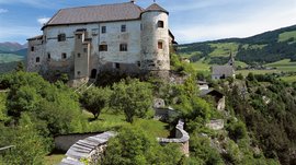 Mächtig und erhaben: Schloss Rodenegg Das Bild zeigt eine alte Burg auf einem grünen Hügel, umgeben von Bäumen und einer Steinmauer. Im Hintergrund ist eine Kirche und eine ländliche Landschaft zu sehen.