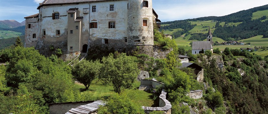 The Gitschberg webcams The picture shows an old castle on a green hill, surrounded by trees and a stone wall. In the background, there is a church and a rural landscape.