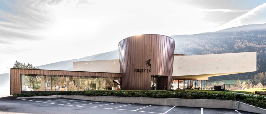 The Gitschberg webcams The picture shows a modern building with a round, copper-colored facade and large glass windows. The building bears the inscription "Capriz," and there is an empty parking lot in front of it, while wooded hills and mountains can be seen in the background.