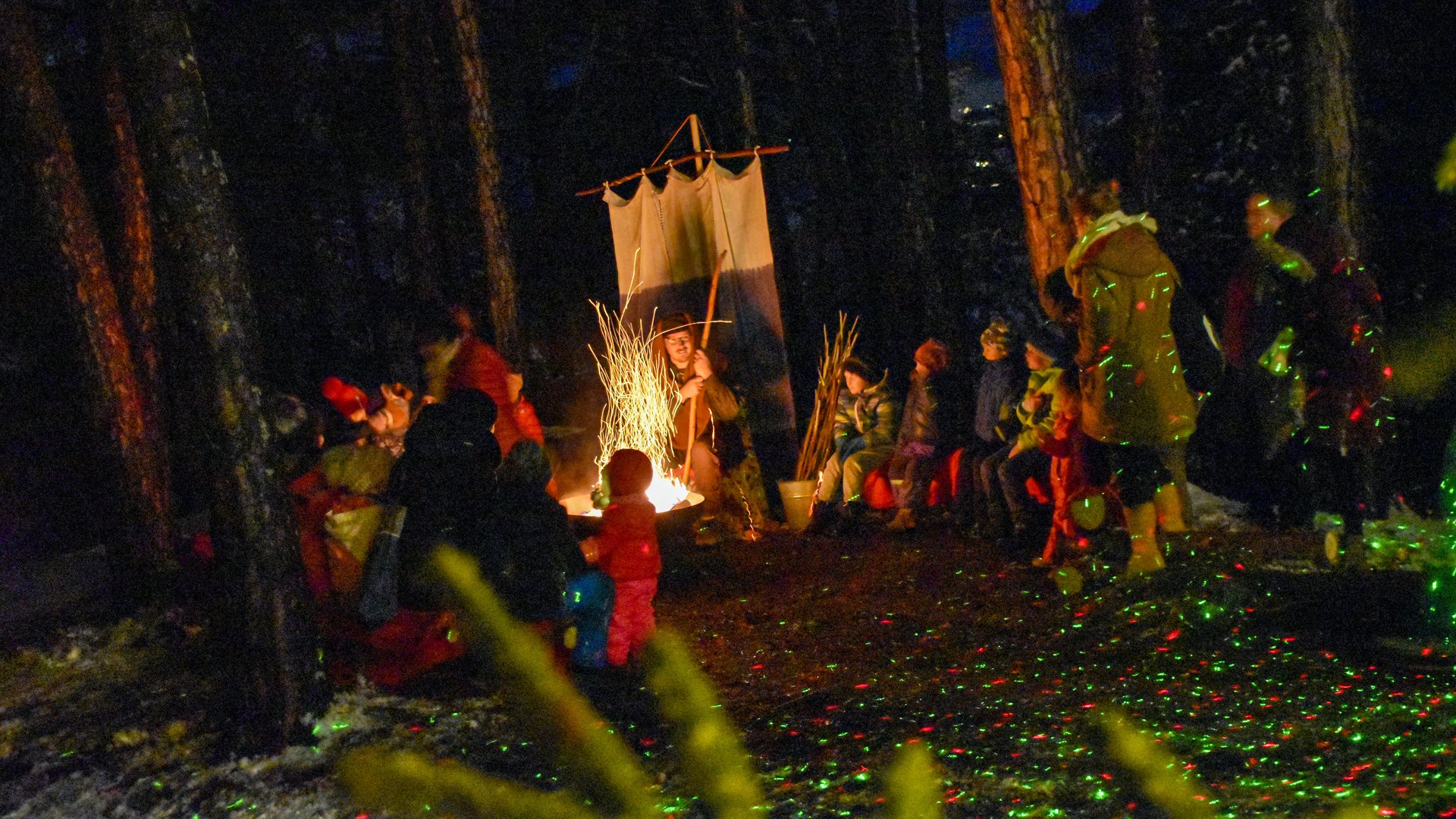 Advent in Terenten The picture shows a group of people sitting around a campfire in a forest. Children and adults are warmly dressed and are listening to a person who is telling stories in the center by the fire. The surroundings are dark, and colorful light spots can be seen on the ground and the trees.