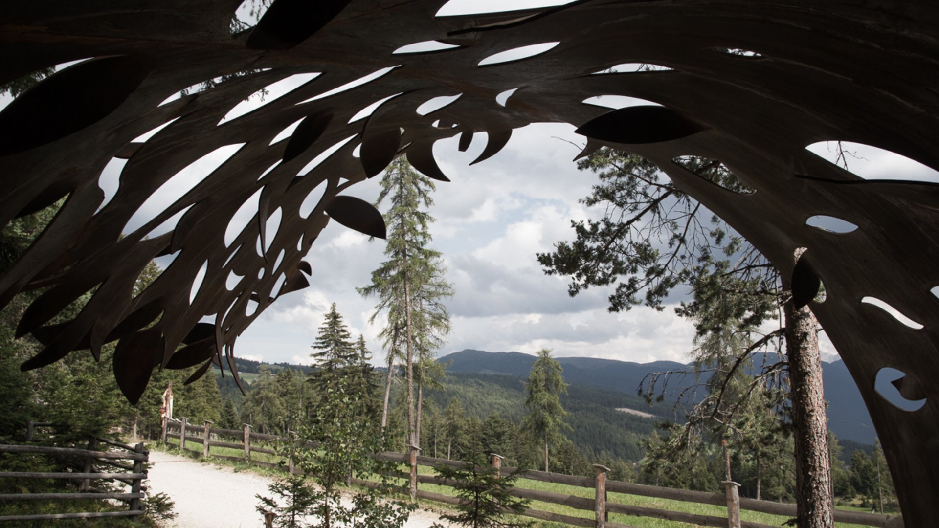 Der Schöpfungsweg in Rodeneck Das Bild zeigt einen Blick durch eine metallische Skulptur mit organischen Formen. Im Hintergrund sind Bäume, ein Weg mit einem Holzzaun und bewaldete Hügel unter einem bewölkten Himmel zu sehen.