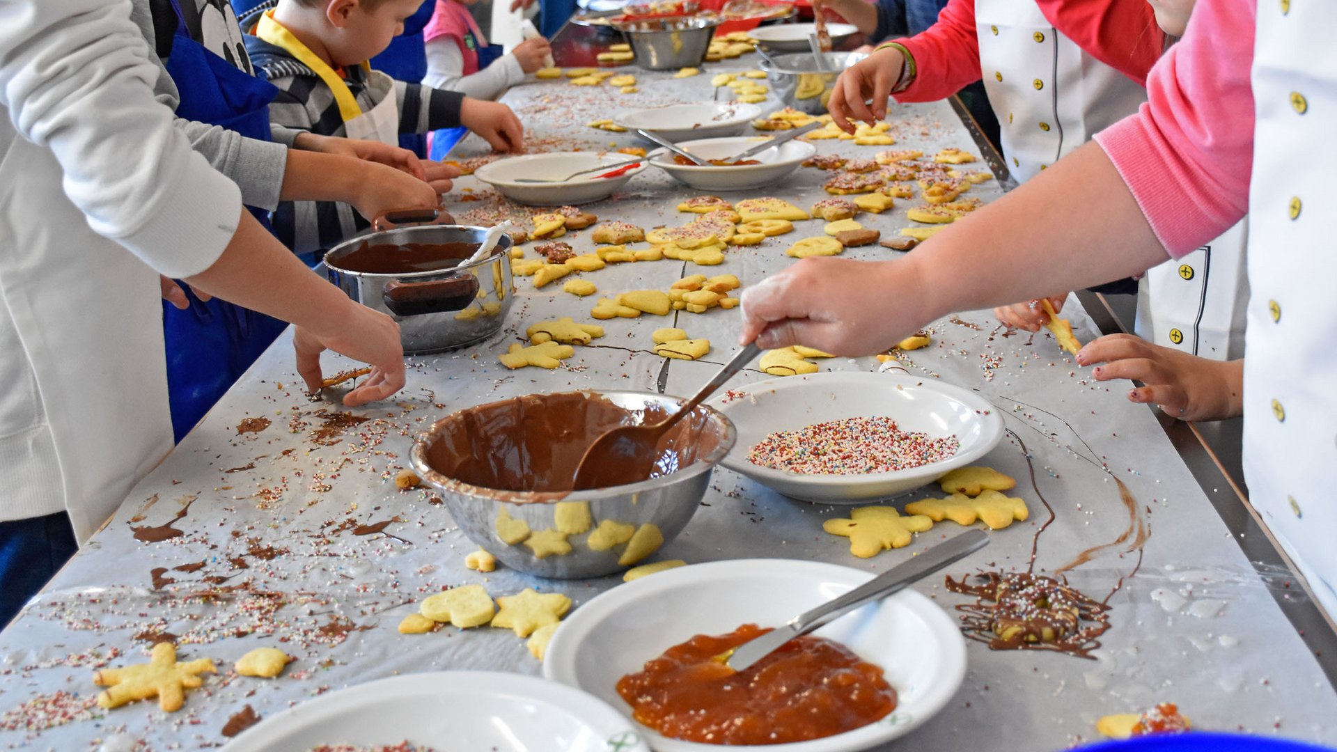 Advent in Terenten The picture shows children decorating cookies. On the table, there are bowls with melted chocolate, jam, and colorful sprinkles, while the children are spreading and decorating the cookies. The table is covered with many finished cookies and ingredients.