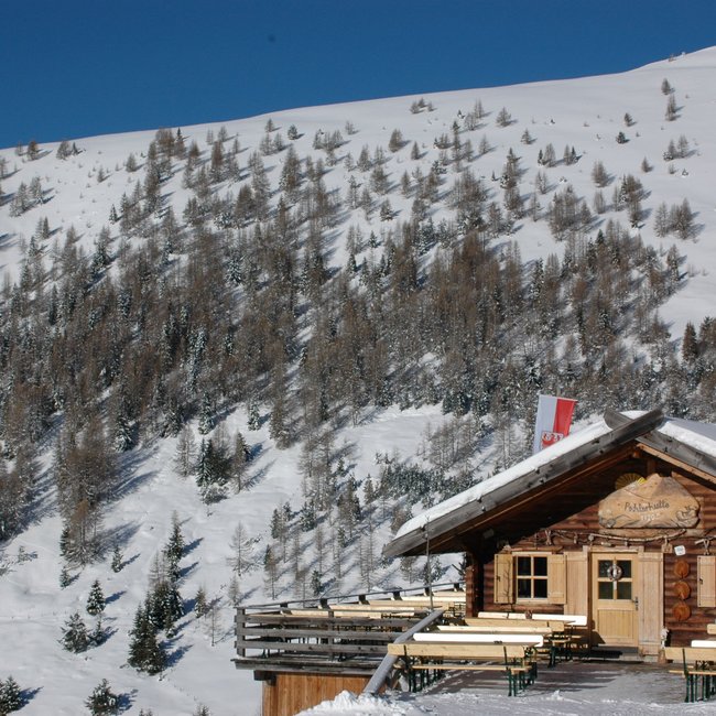 Pichlerhütte De afbeelding toont een knusse houten berghut, omgeven door met sneeuw bedekte hellingen en bomen. Voor de hut staan houten banken en tafels, en boven de ingang hangt een bord met de tekst "Pichlerhütte 1920 m". Naast de hut wappert een rood-wit vlag.