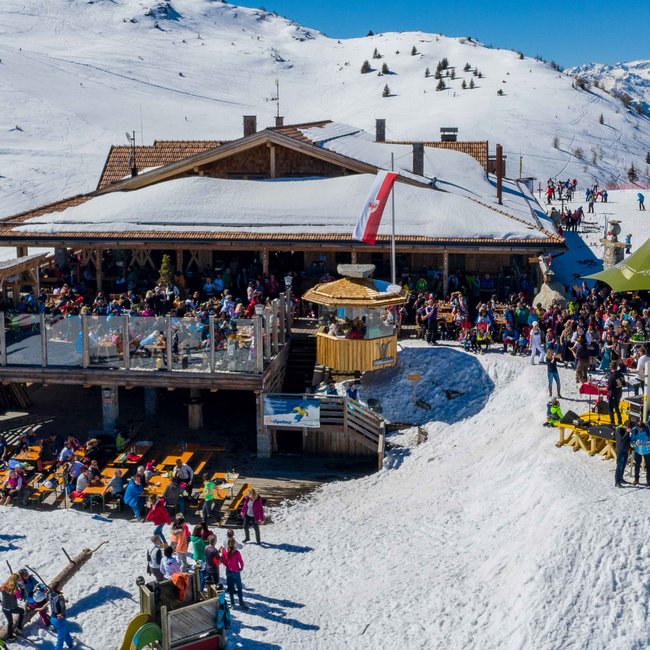 Bergrestaurant Jochtal The picture shows a lively ski lodge with many guests on the terrace, surrounded by snow-covered slopes and skiers, in a sunny alpine winter landscape.