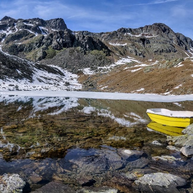Tiefrastenhütte The picture shows a clear mountain lake, partially surrounded by snow and ice, amidst a rocky mountain landscape. A yellow rowboat lies on the shore, and the snow-covered peaks are reflected in the calm water of the lake.