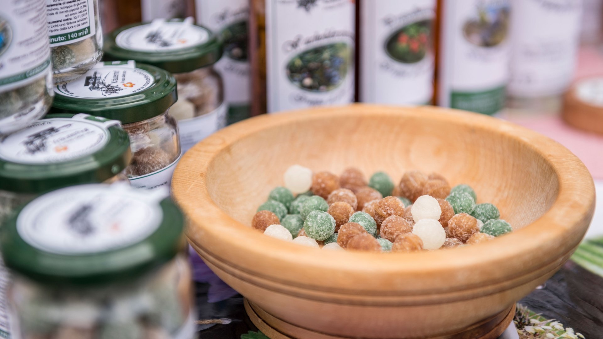 Winkelen in Mühlbach/Italië De afbeelding toont een houten kom gevuld met kleurrijke snoepjes. Op de achtergrond staan verschillende glazen potten met groene deksels, die ook gevuld zijn met snoep of vergelijkbare zoetigheden. Het lijkt op een marktkraam die handgemaakte producten aanbiedt.