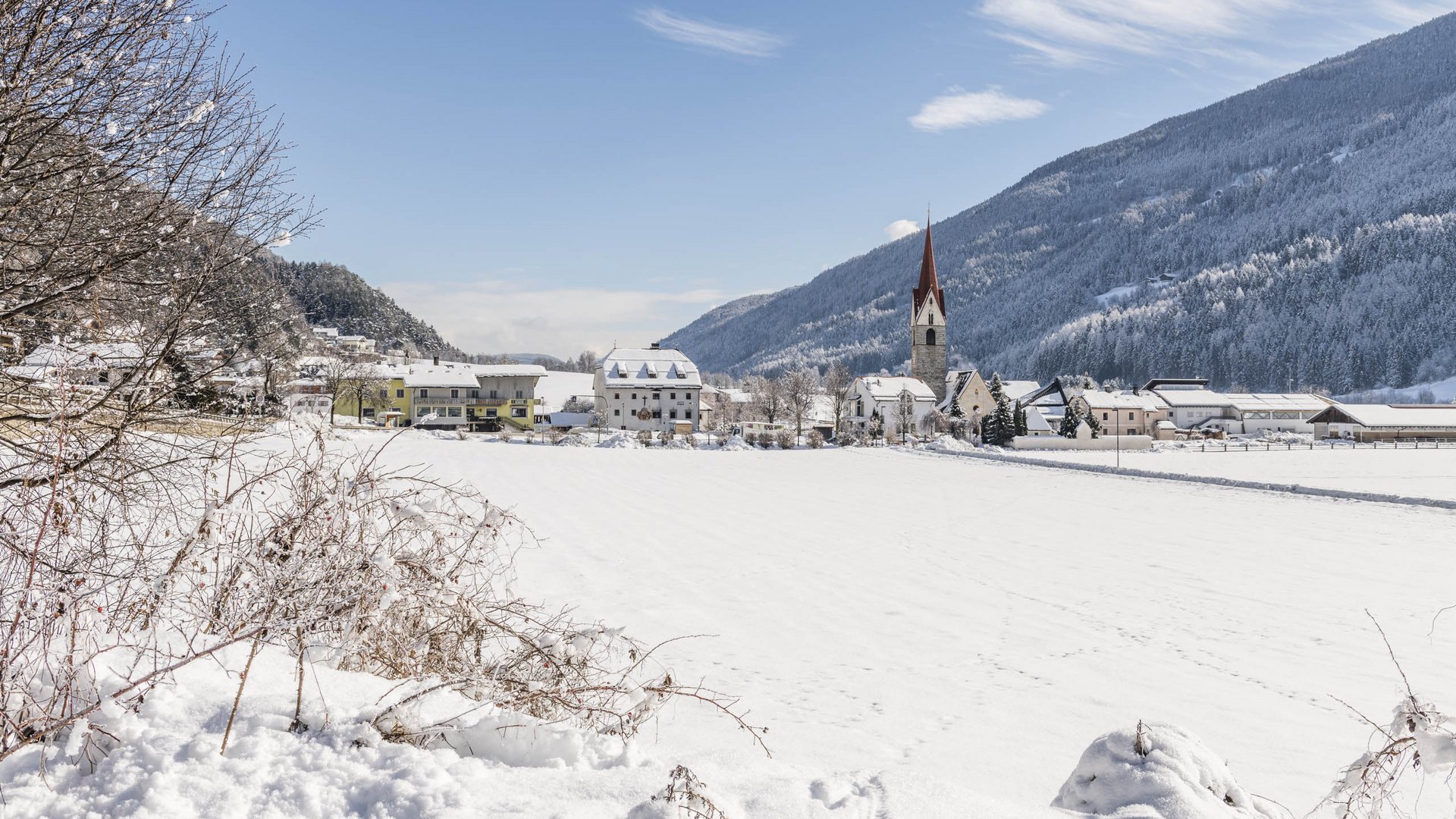 Vintl im Pustertal Das Bild zeigt ein verschneites Dorf, umgeben von schneebedeckten Hügeln und Wäldern. Im Vordergrund steht eine Kirche mit einem spitzen roten Turm, die das Zentrum der ruhigen Winterlandschaft dominiert.