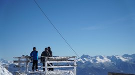 Vals - Jochtal - Steinermandl The picture shows several people in winter clothing on a snow-covered platform. In the background, snow-covered mountains are visible under a bright blue sky.