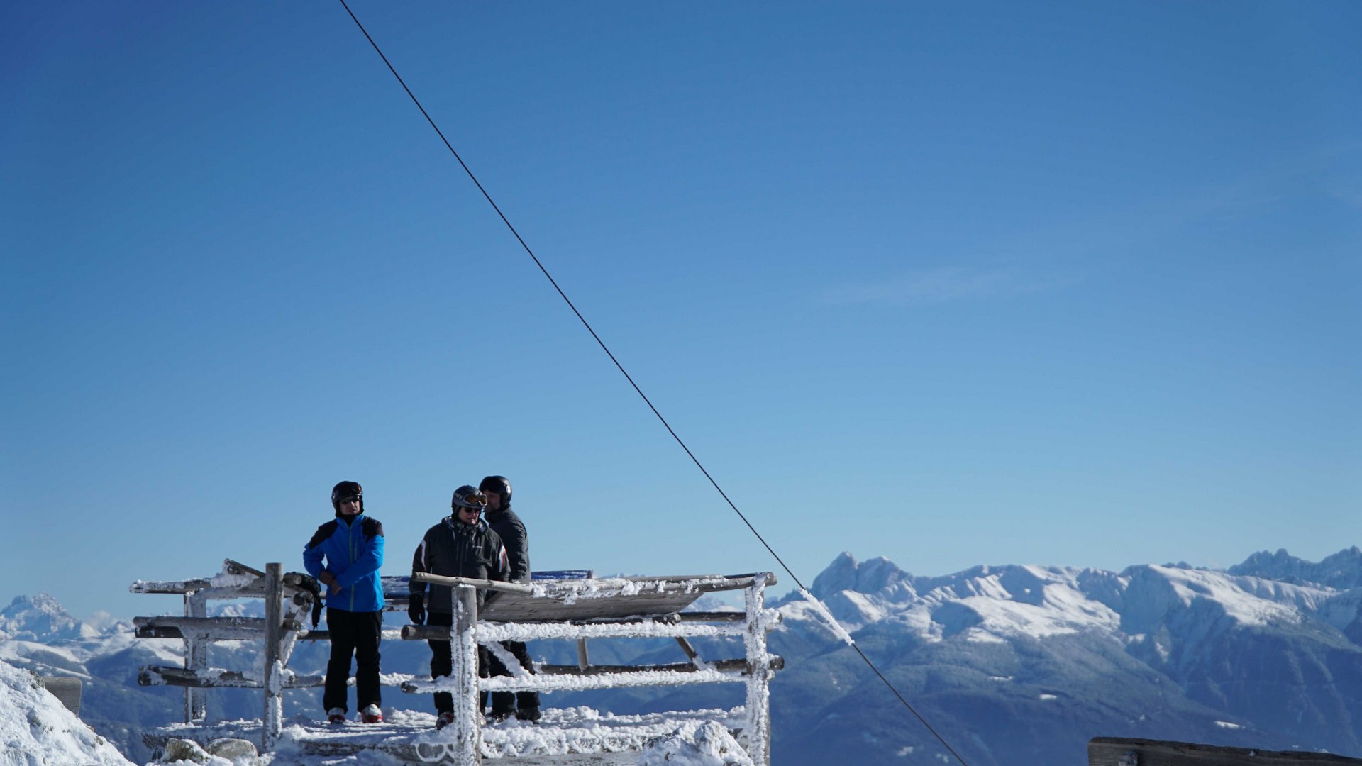 Vals - Jochtal - Steinermandl The picture shows several people in winter clothing on a snow-covered platform. In the background, snow-covered mountains are visible under a bright blue sky.
