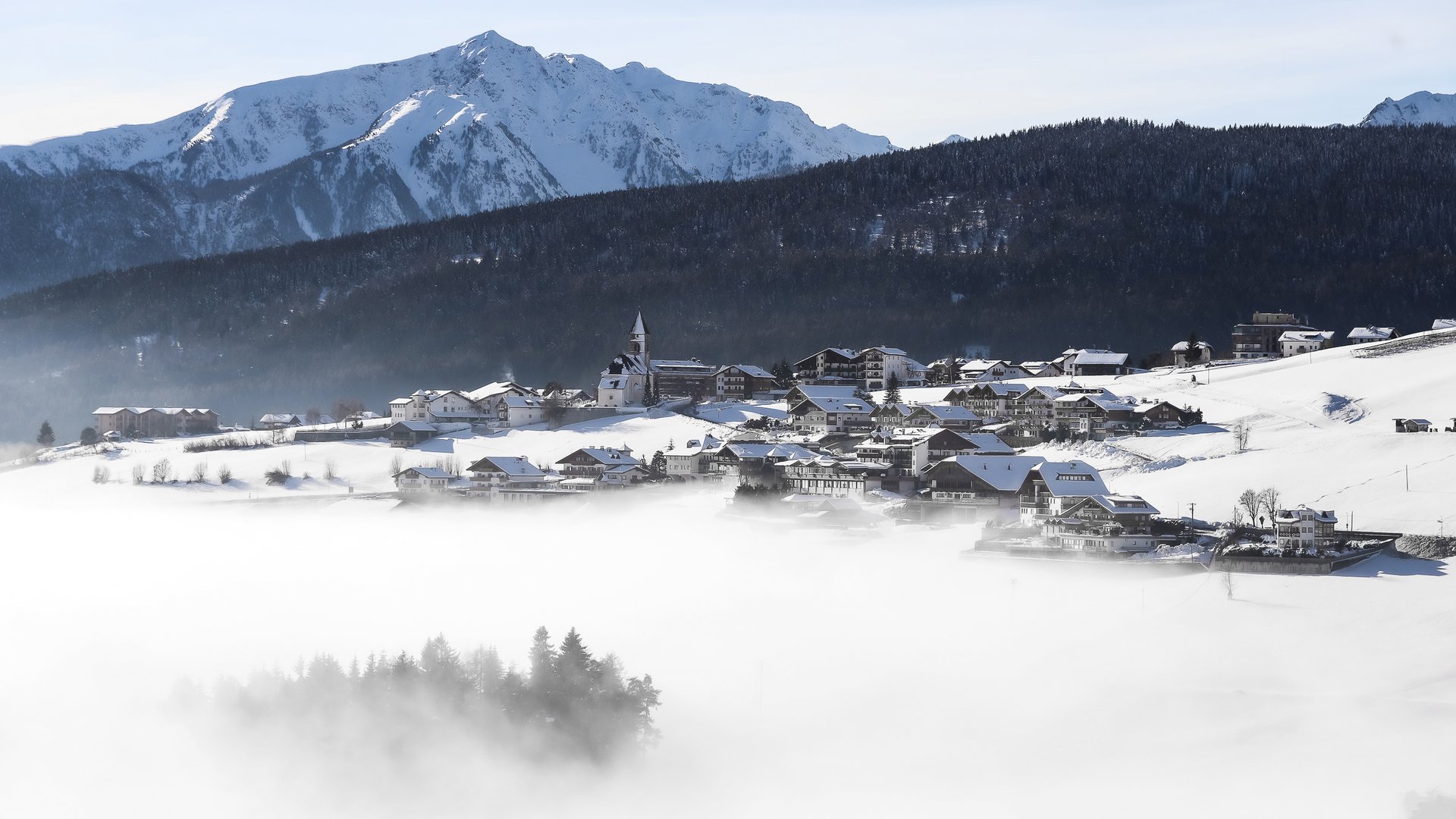 The mountain village of Meransen The picture shows a snow-covered village in a valley surrounded by dense fog. In the background, tall snow-covered mountains and a wooded hillside can be seen. A church stands out in the middle of the village, and the sky is clear and blue.
