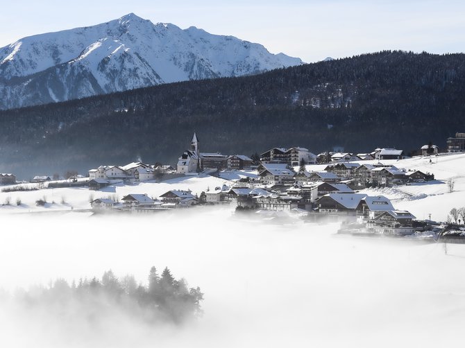 The Alpine hamlet of Spinges The picture shows a snow-covered village in a valley surrounded by dense fog. In the background, tall snow-covered mountains and a wooded hillside can be seen. A church stands out in the middle of the village, and the sky is clear and blue.