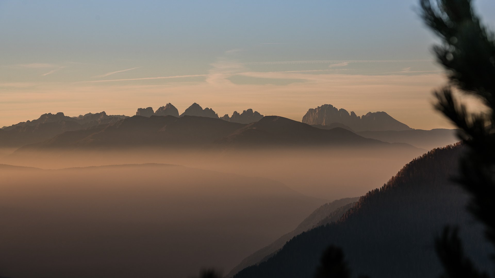 Pfunders: urig und idyllisch Das Bild zeigt eine Berglandschaft bei Sonnenaufgang oder Sonnenuntergang, in der die Silhouetten der Berggipfel gegen den orangefarbenen Himmel hervortreten. Dichte Nebelschichten liegen in den Tälern, was eine mystische Atmosphäre erzeugt.