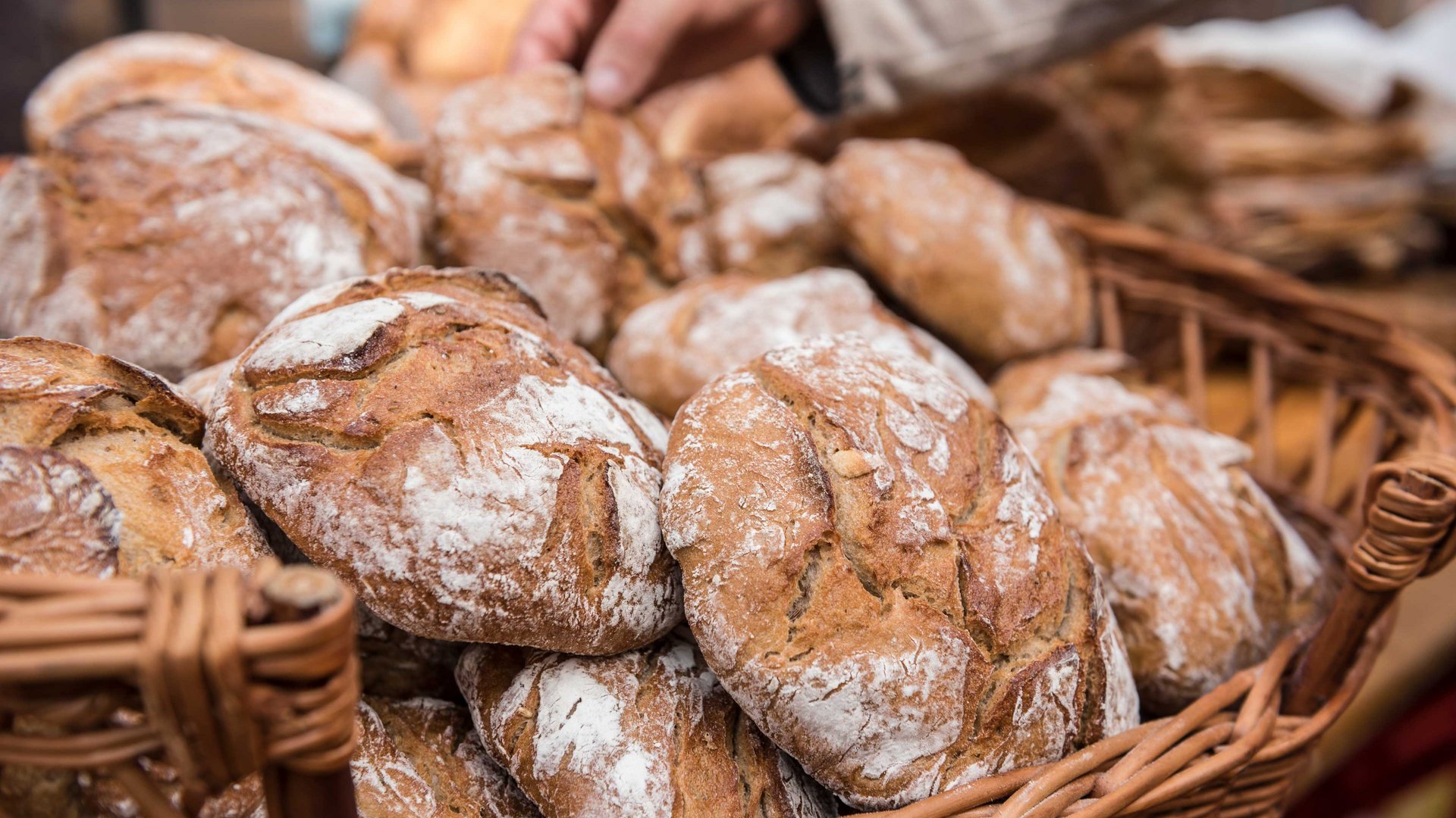 Winkelen in Mühlbach/Italië De afbeelding toont een mand vol versgebakken broden met een knapperige korst en licht bebloemd oppervlak. Een hand grijpt naar een van de broden, wat duidt op een verkoop of een keuze.