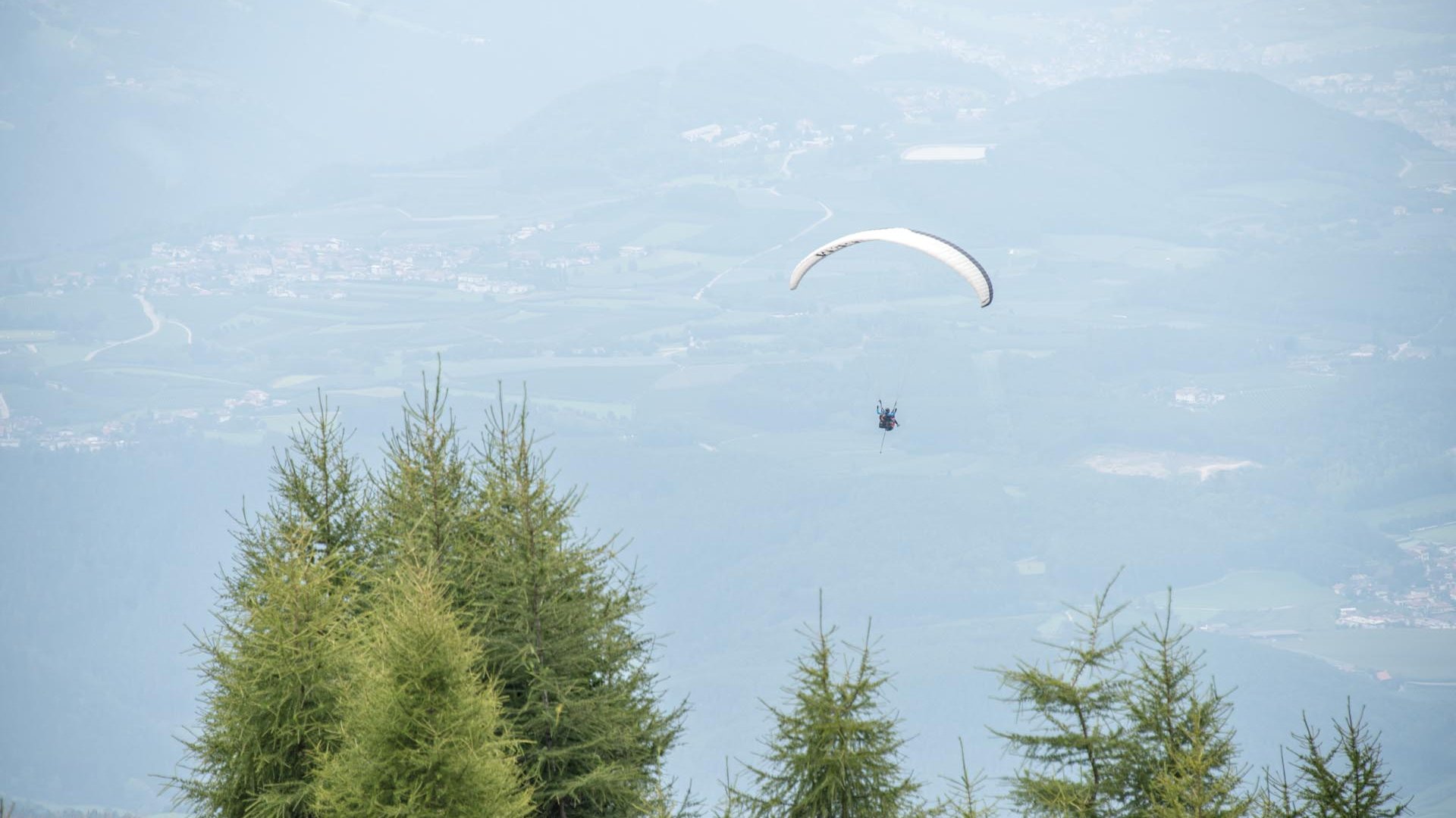 Paragliding v Jižním Tyrolsku Obrázek ukazuje paraglajdistu, který vysoko ve vzduchu nad zalesněnou horou plachtí. V pozadí jsou vidět mírné kopce a údolí s rozesetými vesnicemi a poli.