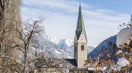 Mühlbach im Pustertal Das Bild zeigt eine Kirche mit einem hohen, grünen Kirchturm in einem verschneiten Dorf. Im Hintergrund sind schneebedeckte Berge und ein blauer Himmel zu sehen, während im Vordergrund kahle Bäume und Sträucher mit Schnee bedeckt sind.