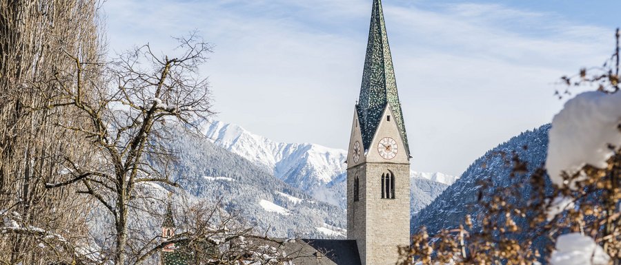 The Gitschberg webcams The picture shows a church with a tall green church tower in a snowy village. In the background, snow-covered mountains and a blue sky are visible, while in the foreground bare trees and bushes are covered with snow.
