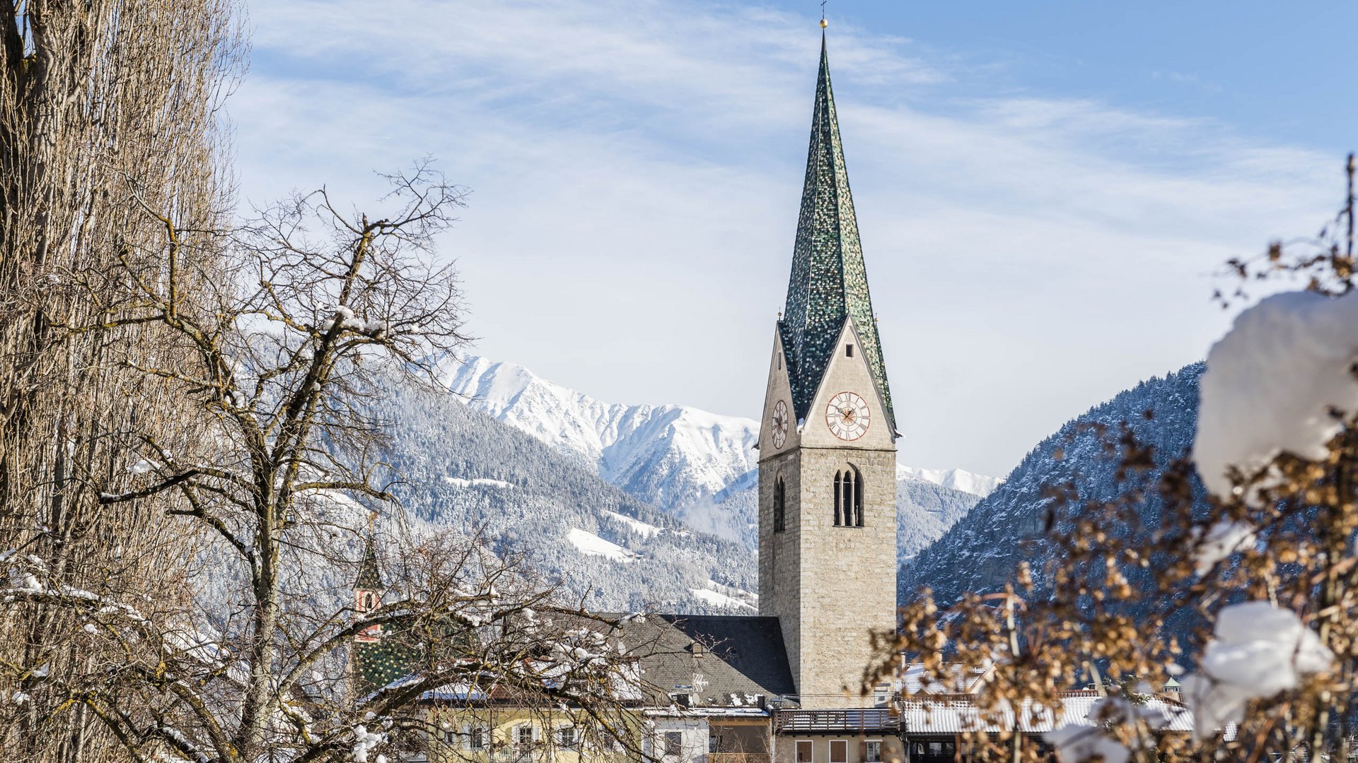 Mühlbach im Pustertal Das Bild zeigt eine Kirche mit einem hohen, grünen Kirchturm in einem verschneiten Dorf. Im Hintergrund sind schneebedeckte Berge und ein blauer Himmel zu sehen, während im Vordergrund kahle Bäume und Sträucher mit Schnee bedeckt sind.