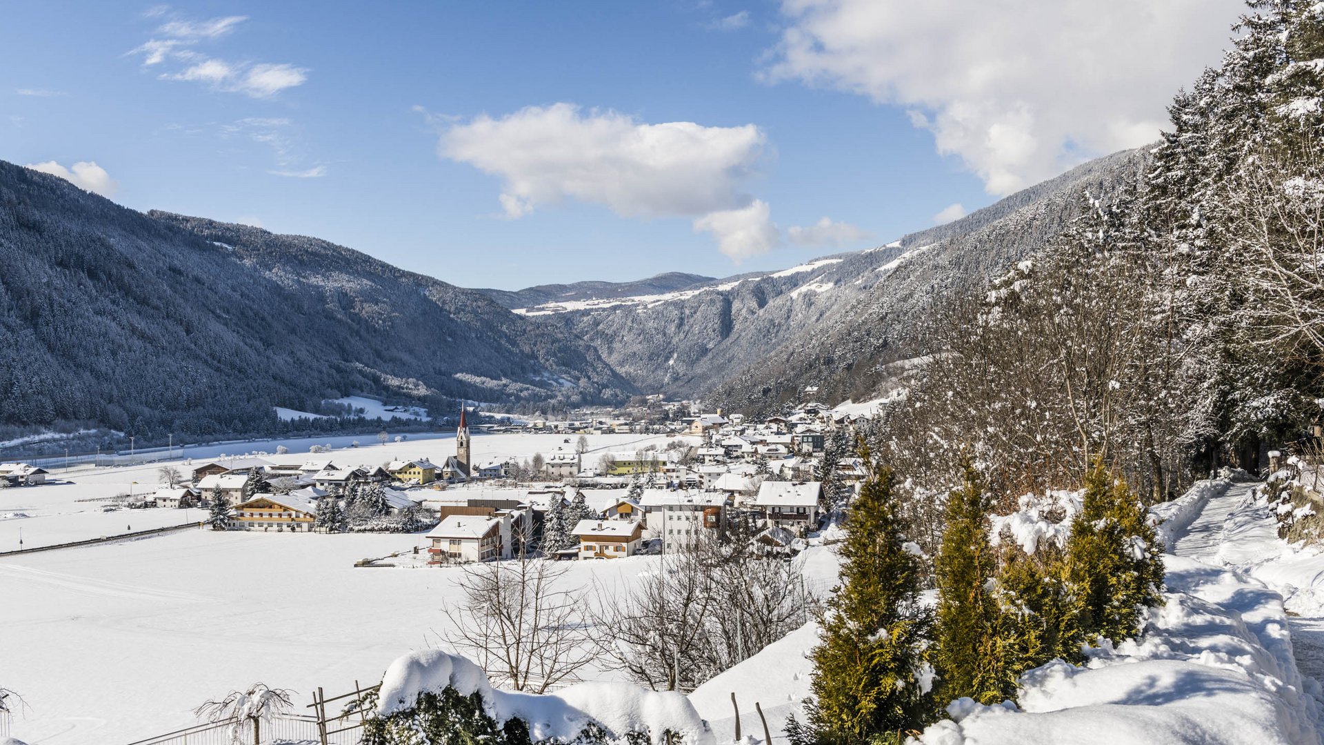 Vintl im Pustertal Das Bild zeigt ein verschneites Dorf in einem weiten Tal, umgeben von schneebedeckten Hügeln und Bergen. Im Vordergrund befindet sich ein schneebedeckter Weg mit Bäumen, und im Hintergrund ist eine Kirche mit einem roten Turm zu sehen, die sich zwischen den Häusern des Dorfes erhebt.