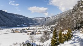 Vintl im Pustertal Das Bild zeigt ein verschneites Dorf in einem weiten Tal, umgeben von schneebedeckten Hügeln und Bergen. Im Vordergrund befindet sich ein schneebedeckter Weg mit Bäumen, und im Hintergrund ist eine Kirche mit einem roten Turm zu sehen, die sich zwischen den Häusern des Dorfes erhebt.