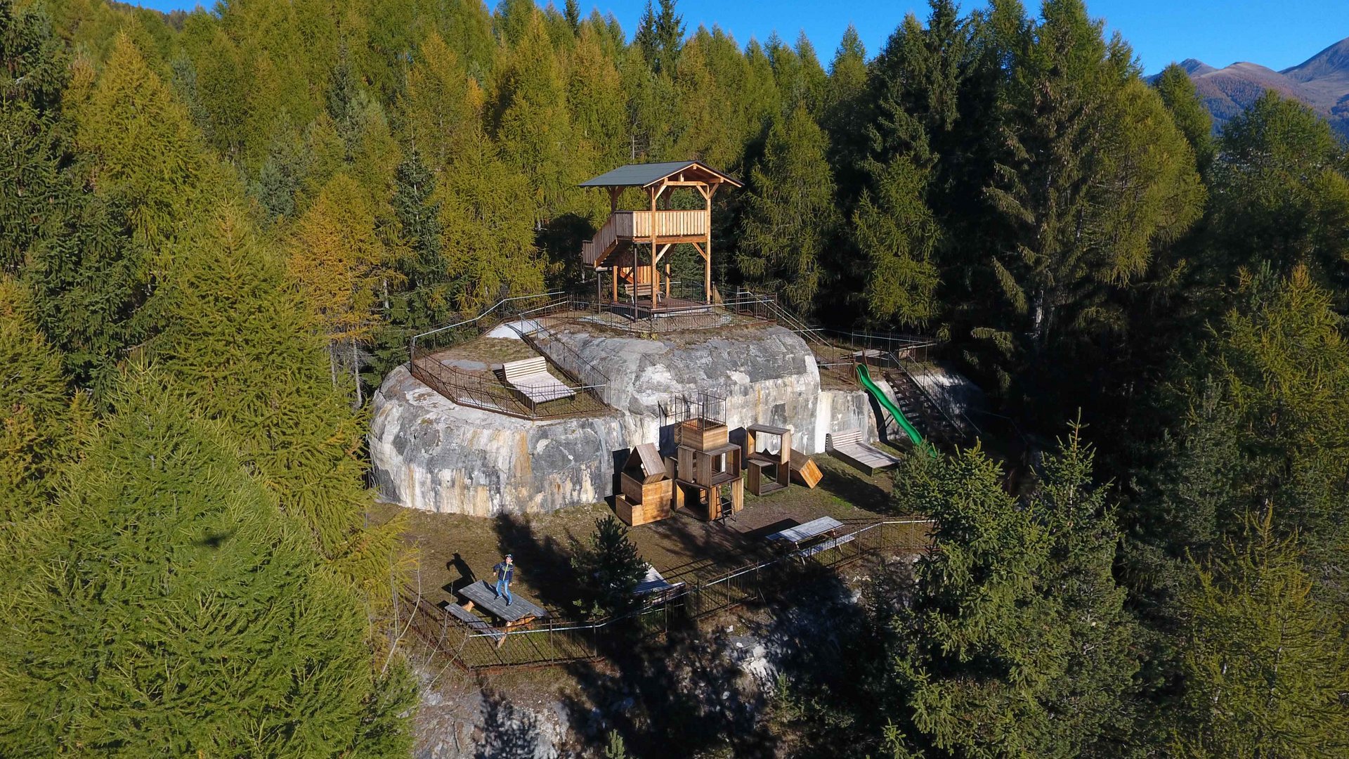 The recreation area in Spinges, South Tyrol The image shows a rocky hill in a forested area, on which a wooden structure resembling a lookout point or platform has been erected. In the foreground there are seating options, playground equipment, and a slide tower, surrounded by dense green trees and a clear, blue sky in the background.