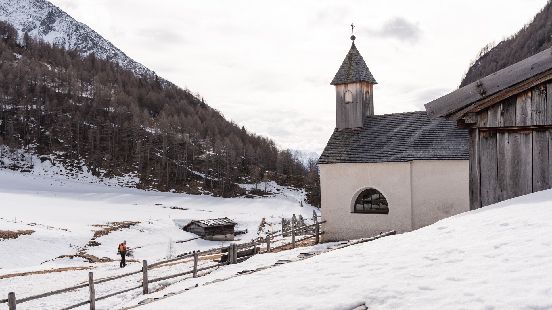 Fane Alm – South Tyrol’s most idyllic Alpine village The picture shows a snowy landscape with a small, simple chapel that has a wooden shingle roof and a tower with a cross. In the background, snow-covered hills and trees are visible, while a hiker walks along a path next to a wooden fence.