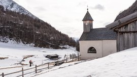 Fane Alm – South Tyrol’s most idyllic Alpine village The picture shows a snowy landscape with a small, simple chapel that has a wooden shingle roof and a tower with a cross. In the background, snow-covered hills and trees are visible, while a hiker walks along a path next to a wooden fence.