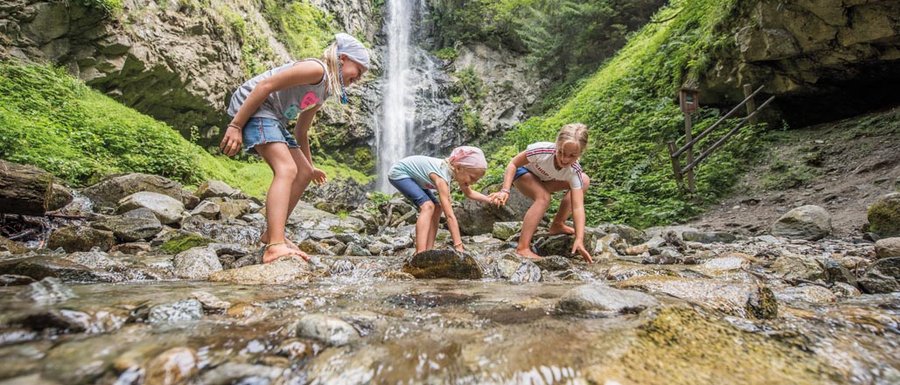 The Gitschberg webcams The picture shows three children playing barefoot in a shallow stream. In the background, there is a large waterfall cascading down a steep rock face, surrounded by lush, green forest.