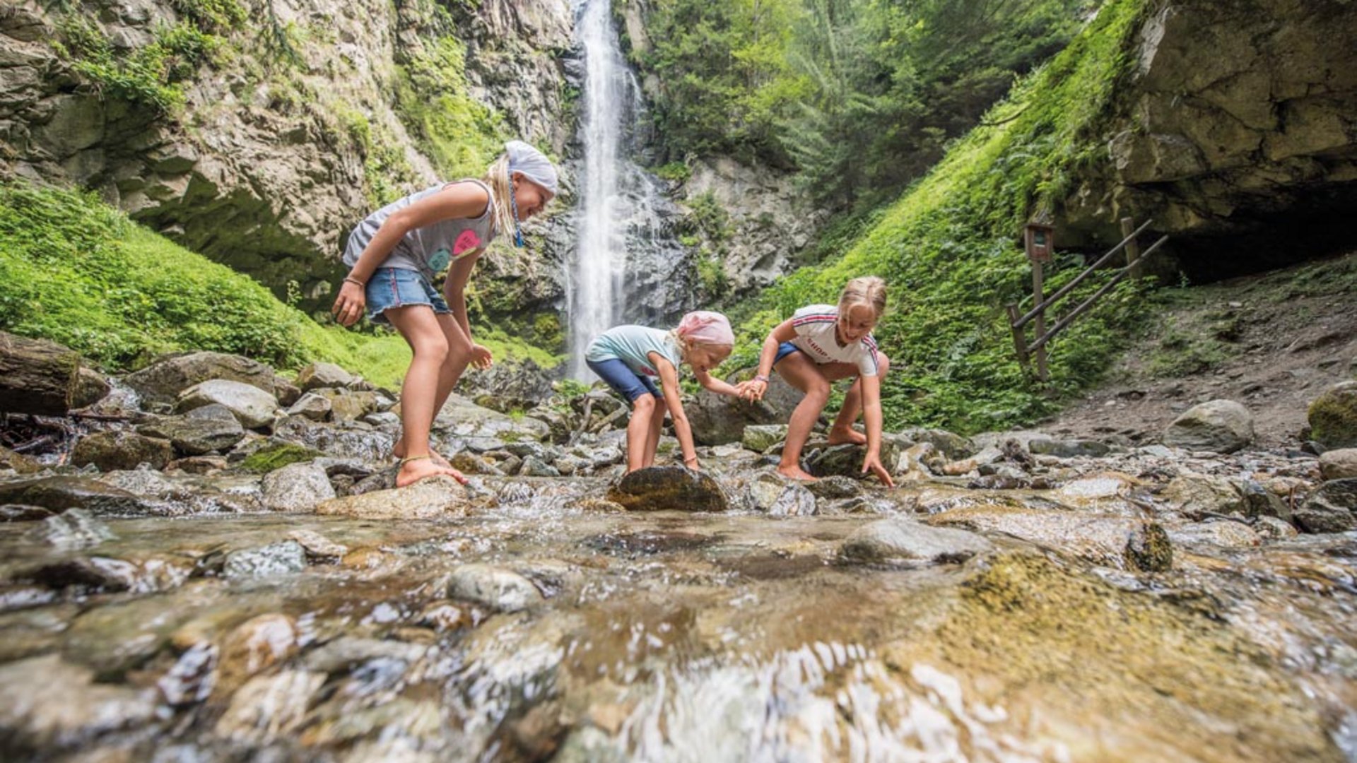 De Elfenweg bij Vintl De afbeelding toont drie kinderen die blootsvoets in een ondiepe beek spelen. Op de achtergrond is een grote waterval te zien die van een steile rotswand naar beneden stort, omgeven door een weelderig, groen bos.