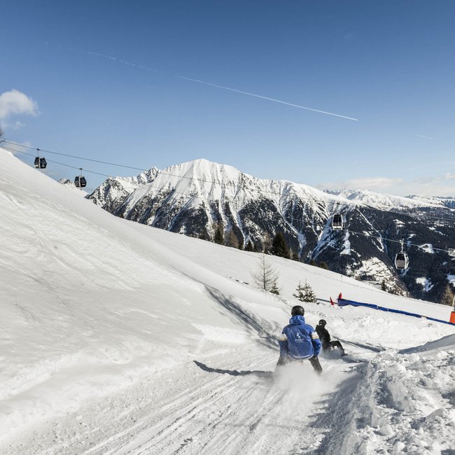 Rodeln in Südtirol Personen rodeln auf verschneiter Piste mit Seilbahn und Bergen im Hintergrund