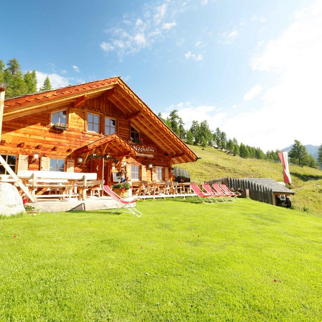 Nockalm Obrázek ukazuje útulnou dřevěnou chatu nazvanou "Nockalm" na zelené louce, obklopenou lehátky a stoly. V pozadí jsou vidět zalesněné kopce a jasná modrá obloha.