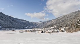 Vintl im Pustertal Das Bild zeigt ein verschneites Tal, in dem ein kleines Dorf liegt, umgeben von bewaldeten, schneebedeckten Bergen. Der Himmel ist blau mit einigen Wolken, und die verschneite Landschaft wirkt ruhig und friedlich.