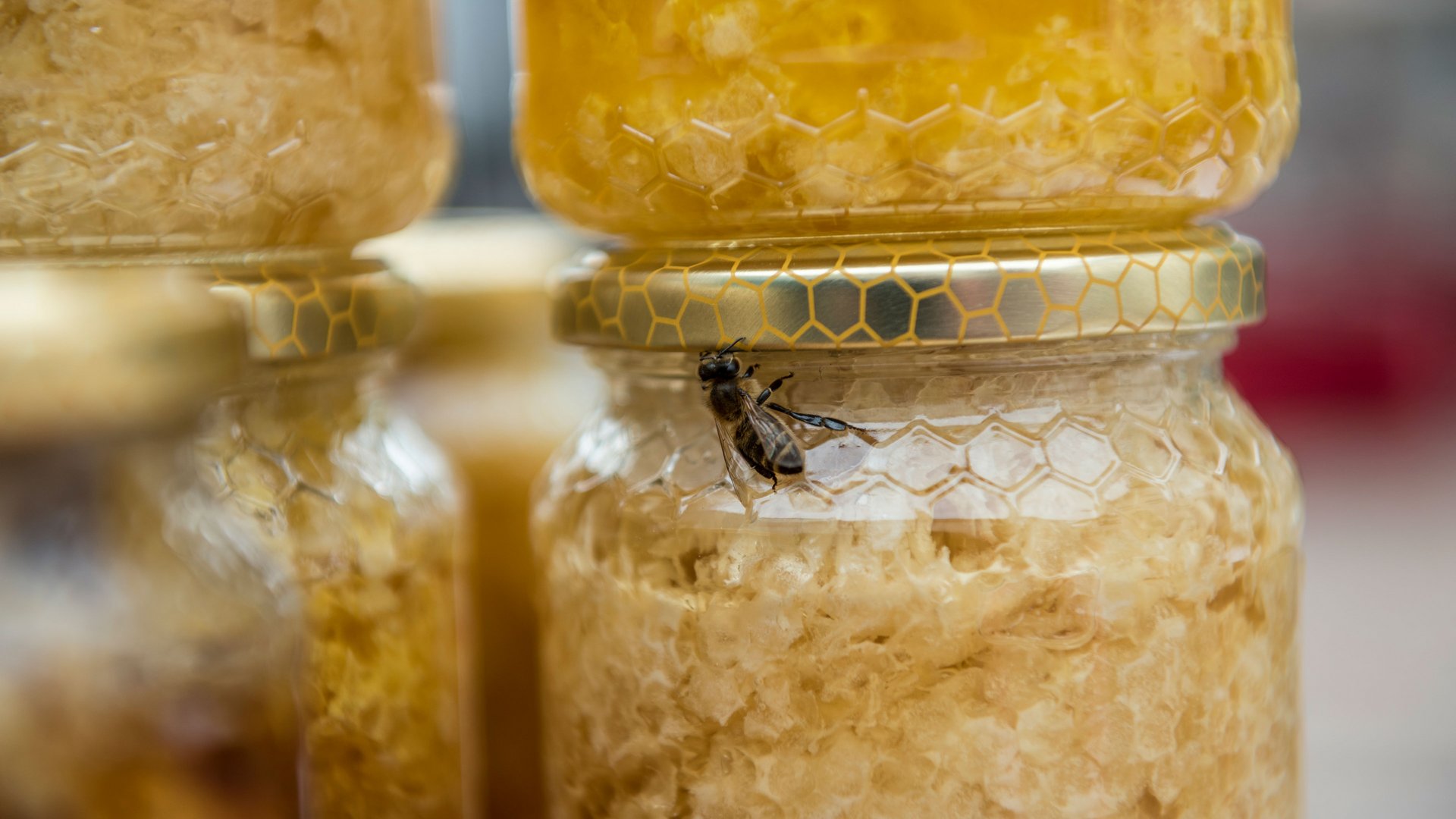 Winkelen in Mühlbach/Italië De afbeelding toont een pot met honingraten, op het deksel ervan zit een bij. De pot heeft een gouden etiket met honingraatpatroon, en op de achtergrond zijn meer honingpotten te zien.
