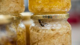 Winkelen in Mühlbach/Italië De afbeelding toont een pot met honingraten, op het deksel ervan zit een bij. De pot heeft een gouden etiket met honingraatpatroon, en op de achtergrond zijn meer honingpotten te zien.