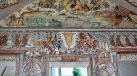 Mächtig und erhaben: Schloss Rodenegg Das Bild zeigt ein detailliertes Wandfresko in einem historischen Raum, das religiöse und adelige Figuren sowie Wappen darstellt. Die Szenen sind farbenfroh und reich verziert.