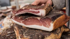 Winkelen in Mühlbach/Italië De afbeelding toont een hand die een groot stuk spek vasthoudt. Het spek heeft een donker gekruide korst en toont duidelijk de witte vetlagen en het donkerrode vlees aan de binnenkant. Meerdere stukken spek liggen opgestapeld op een ondergrond.