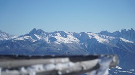 Vals - Jochtal - Steinermandl The picture shows a vast mountain landscape with snow-covered peaks under a clear blue sky. In the foreground, a blurry wooden railing is visible, directing the view toward the majestic mountains in the background.