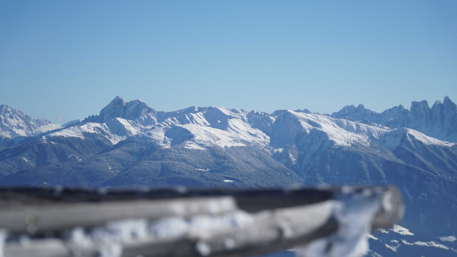 Vals - Jochtal - Steinermandl The picture shows a vast mountain landscape with snow-covered peaks under a clear blue sky. In the foreground, a blurry wooden railing is visible, directing the view toward the majestic mountains in the background.