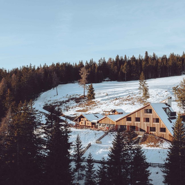 Oberhauserhütte Das Bild zeigt eine moderne Berghütte aus Holz, die von schneebedeckten Hügeln und dichtem Wald umgeben ist. Die Hütte liegt in einer malerischen Winterlandschaft mit Tannenbäumen im Vordergrund und einem klaren, blauen Himmel darüber.