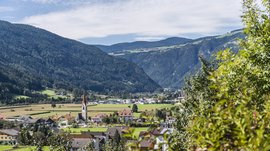 Vintl im Pustertal Das Bild zeigt ein Tal mit einem kleinen Dorf, das von bewaldeten Hügeln umgeben ist. In der Mitte des Dorfs ragt ein Kirchturm mit rotem Dach hervor, während sich im Hintergrund grüne Felder und Wälder bis zu den Hügeln erstrecken.