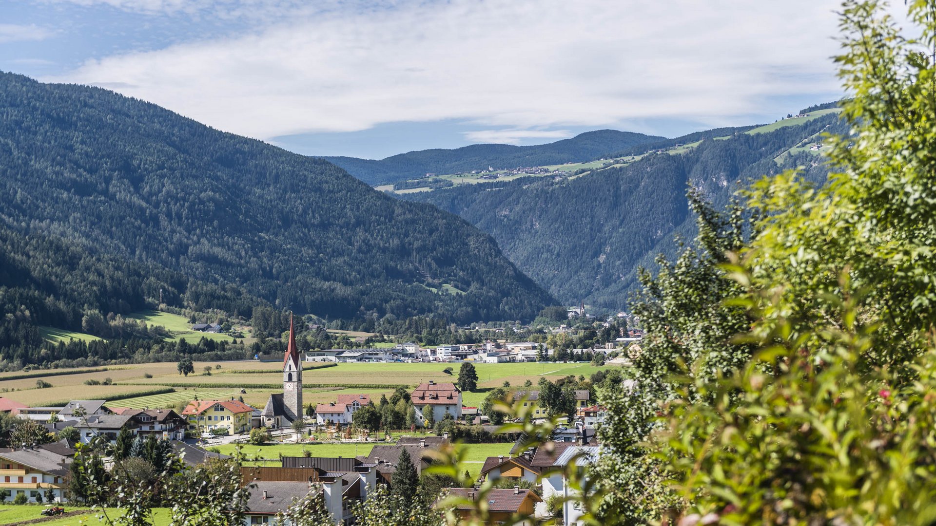 Vintl im Pustertal Das Bild zeigt ein Tal mit einem kleinen Dorf, das von bewaldeten Hügeln umgeben ist. In der Mitte des Dorfs ragt ein Kirchturm mit rotem Dach hervor, während sich im Hintergrund grüne Felder und Wälder bis zu den Hügeln erstrecken.
