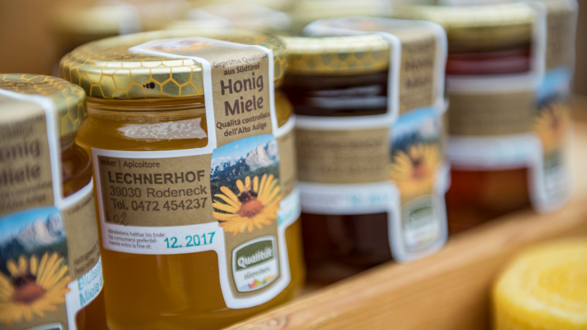 Winkelen in Mühlbach/Italië De afbeelding toont meerdere potten honing die op een plank staan. Op de etiketten van de potten staat "Honig Miele" evenals het adres en contactgegevens van een imker die werkzaam is op een boerderij genaamd "Lechnerhof" in Rodeneck, Zuid-Tirol. De potten hebben goudkleurige deksels met een honingraatpatroon.