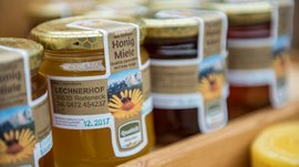 Winkelen in Mühlbach/Italië De afbeelding toont meerdere potten honing die op een plank staan. Op de etiketten van de potten staat "Honig Miele" evenals het adres en contactgegevens van een imker die werkzaam is op een boerderij genaamd "Lechnerhof" in Rodeneck, Zuid-Tirol. De potten hebben goudkleurige deksels met een honingraatpatroon.