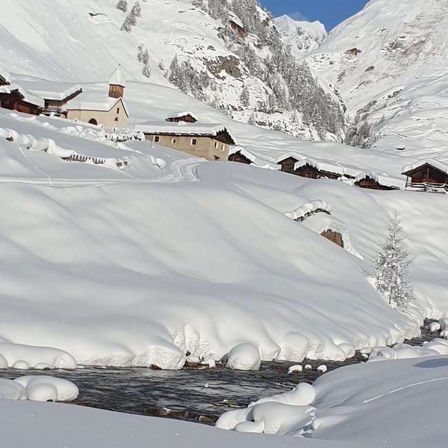 Zingerle Hütte De afbeelding toont een diep met sneeuw bedekt berglandschap met een kleine verzameling hutten en een kerk in het dal. Op de voorgrond stroomt een klein beekje door het besneeuwde landschap, en op de achtergrond zijn hoge, met sneeuw bedekte bergen te zien onder een heldere, blauwe hemel.