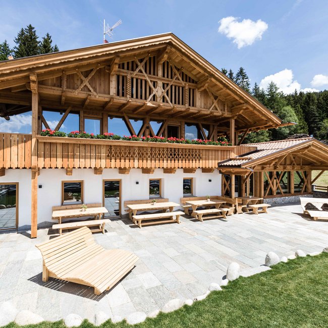 Ahner Berghof Obrázek ukazuje elegantní, tradiční alpský dům s dřevěnou fasádou a balkonem, zdobeným červenými květinami, na pozadí zelených stromů a dobře udržované kamenné desky s několika dřevěnými stoly a venkovní lavicí.