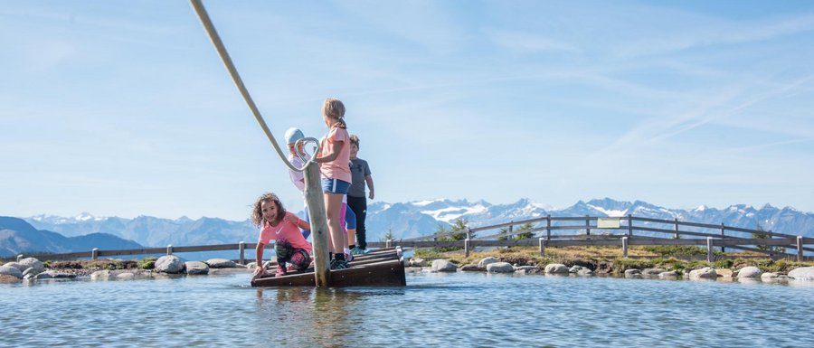 The Gitschberg webcams The picture shows several children standing on a small raft, which is being pulled with a rope across a shallow pond. In the background, there are mountains that complement the clear blue sky and the picturesque landscape.